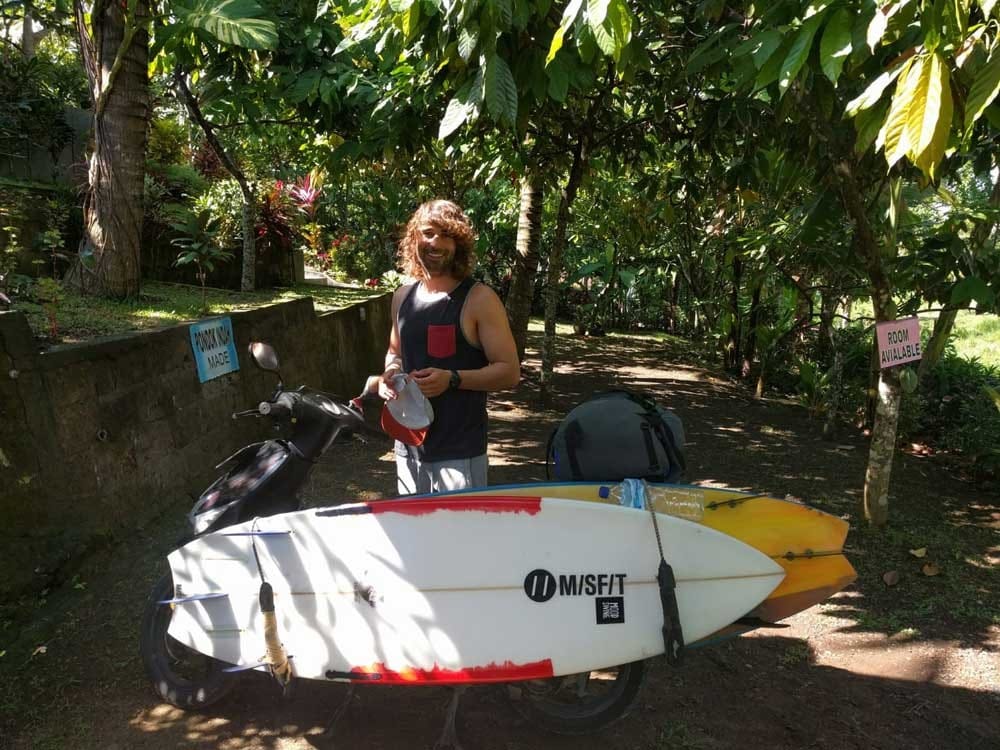 estudiante Nueva Zelanda chico con el pelo largo y una tabla de surf sonriendo a cámara y está entre la vegetación